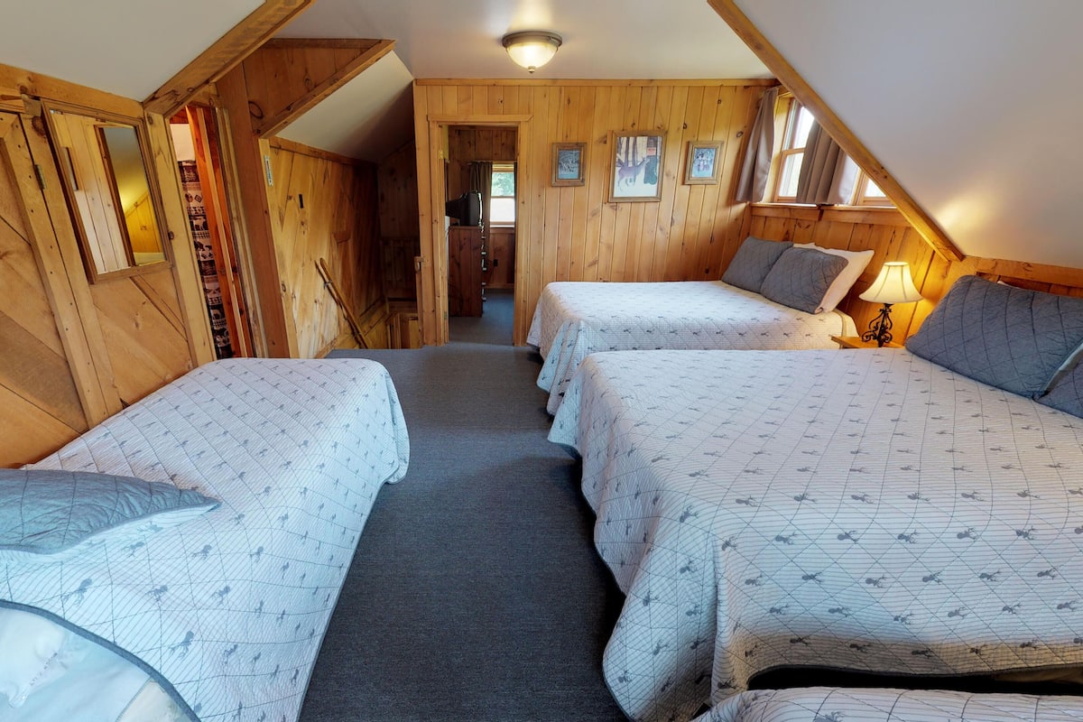 A room with wooden walls and a sloped ceiling, featuring two beds with patterned bedding and a small table with a lamp.