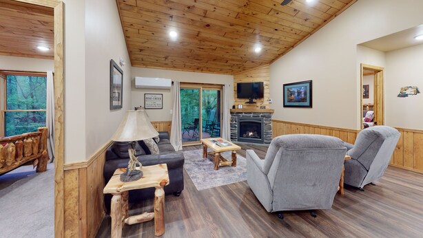 A cozy living room with a stone fireplace, wooden furniture, and a large window offering a view of the forest.