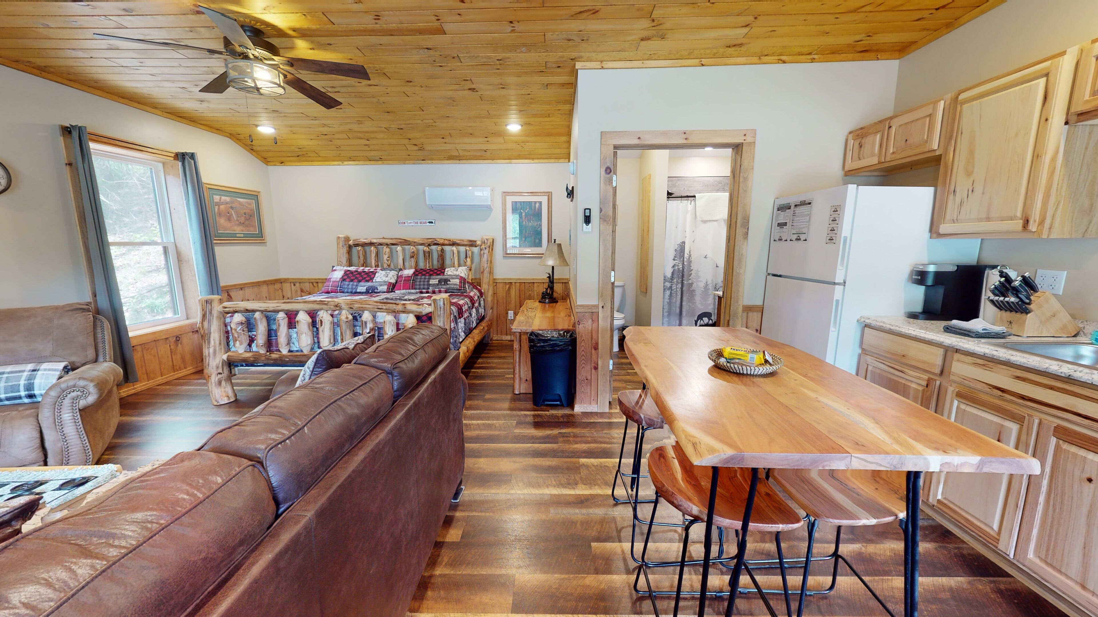 Interior acogedor de una cabaña con techo de madera, una larga mesa de comedor con taburetes altos, un sofá y una cama con cabecera de madera.