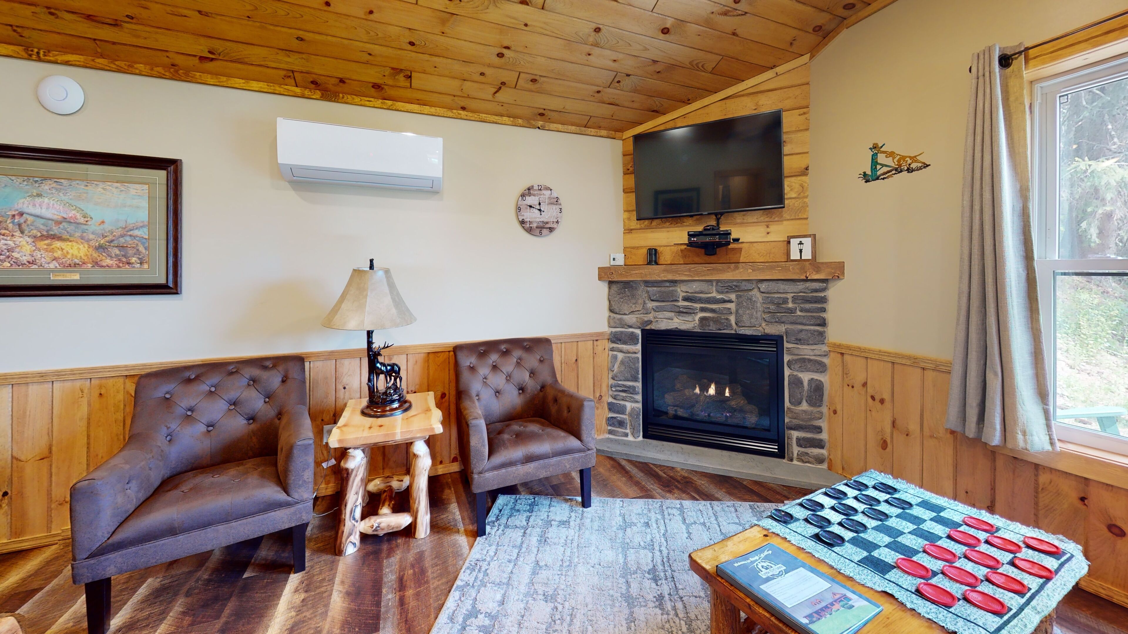 A cozy living room with a fireplace, two leather armchairs, a table with a chessboard, and a television mounted above the fireplace.