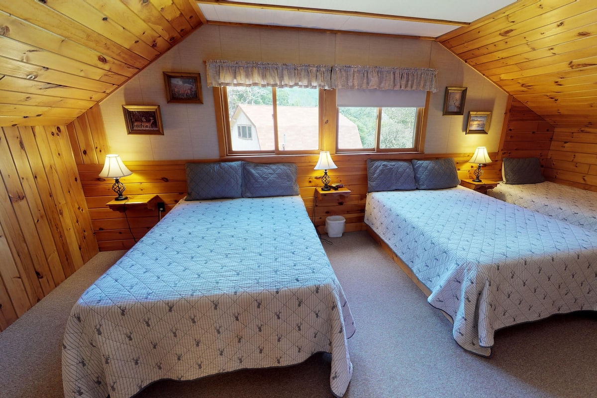 A cozy attic bedroom with two beds, wooden walls, a window with a view, and a small window with curtains.