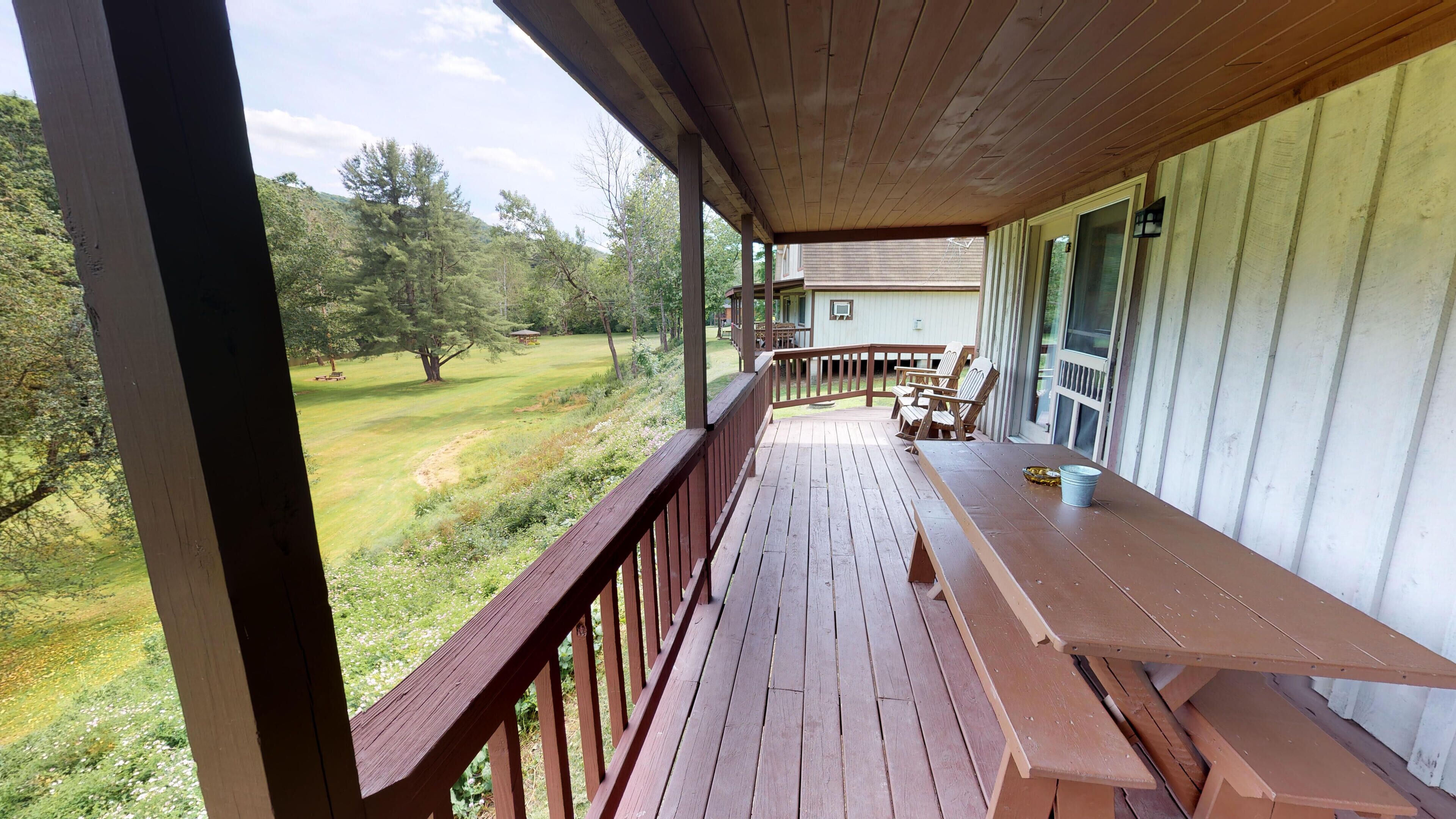 A wooden deck with a bench and chairs, overlooking a grassy yard with trees.
