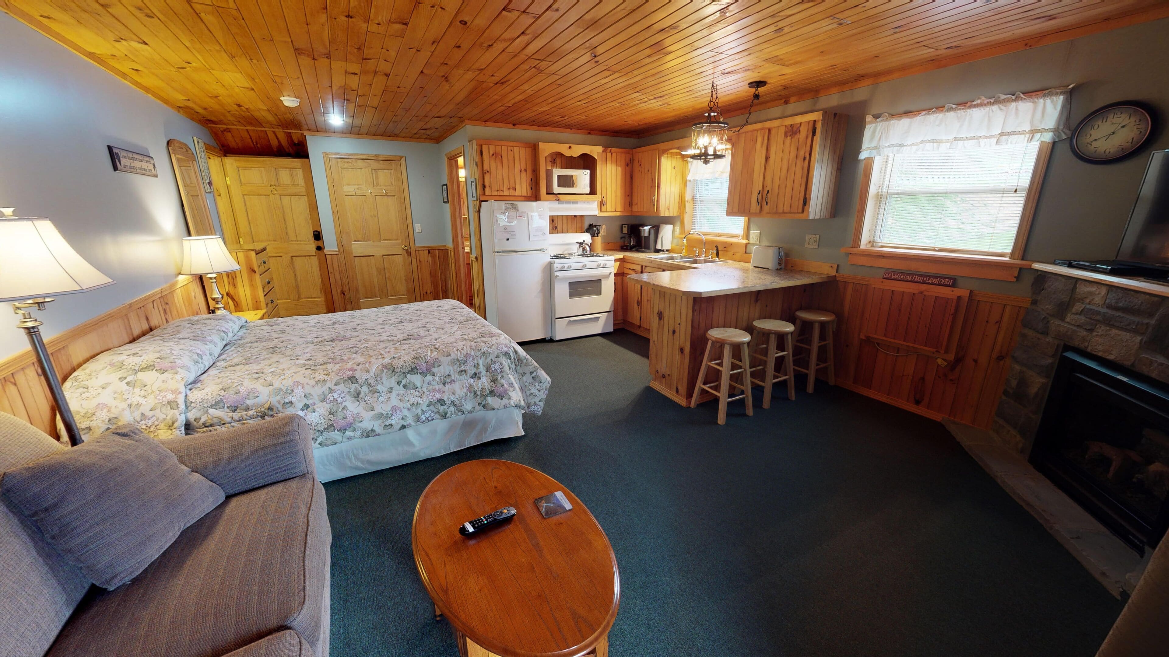 A cozy cabin with a bed, a small table, a kitchen with wooden cabinets, a stove, and a fireplace.