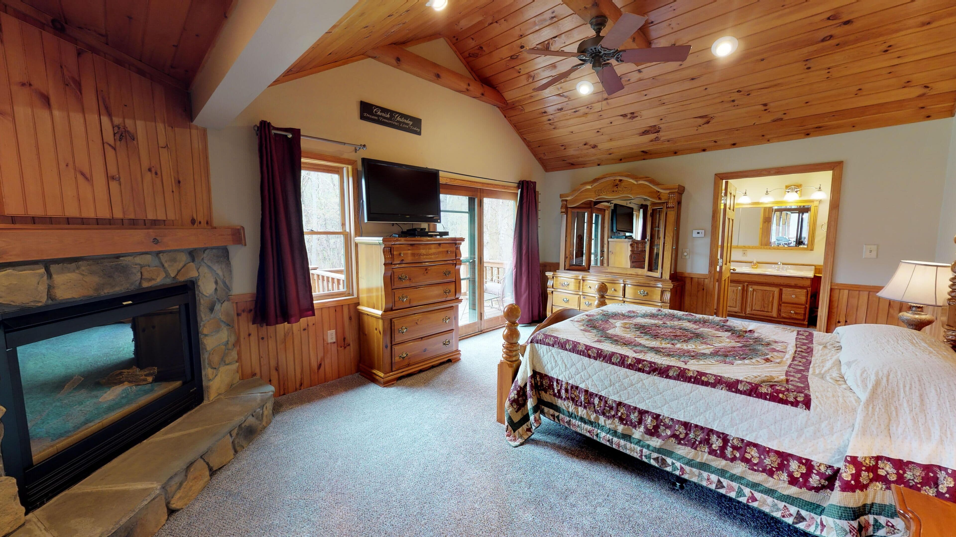 A cozy bedroom with a stone fireplace, wooden ceiling, a large bed with a floral quilt, a wooden dresser, a television, and a large mirror.