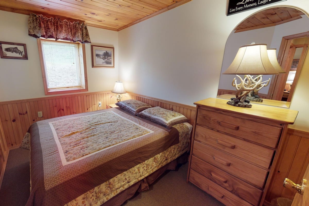 A bedroom with a wooden bed, a nightstand with a lamp, a dresser, and a window with curtains.