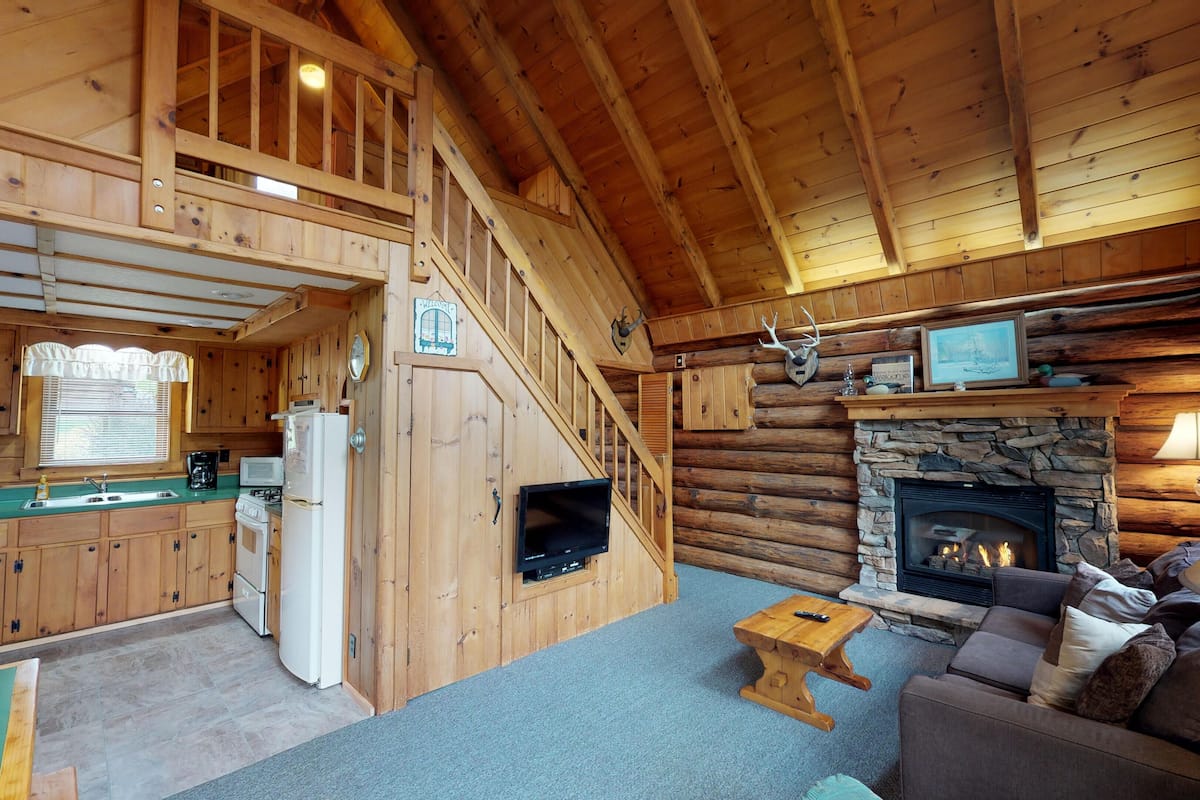 A cabin interior with a kitchen, living room, fireplace, and a staircase.