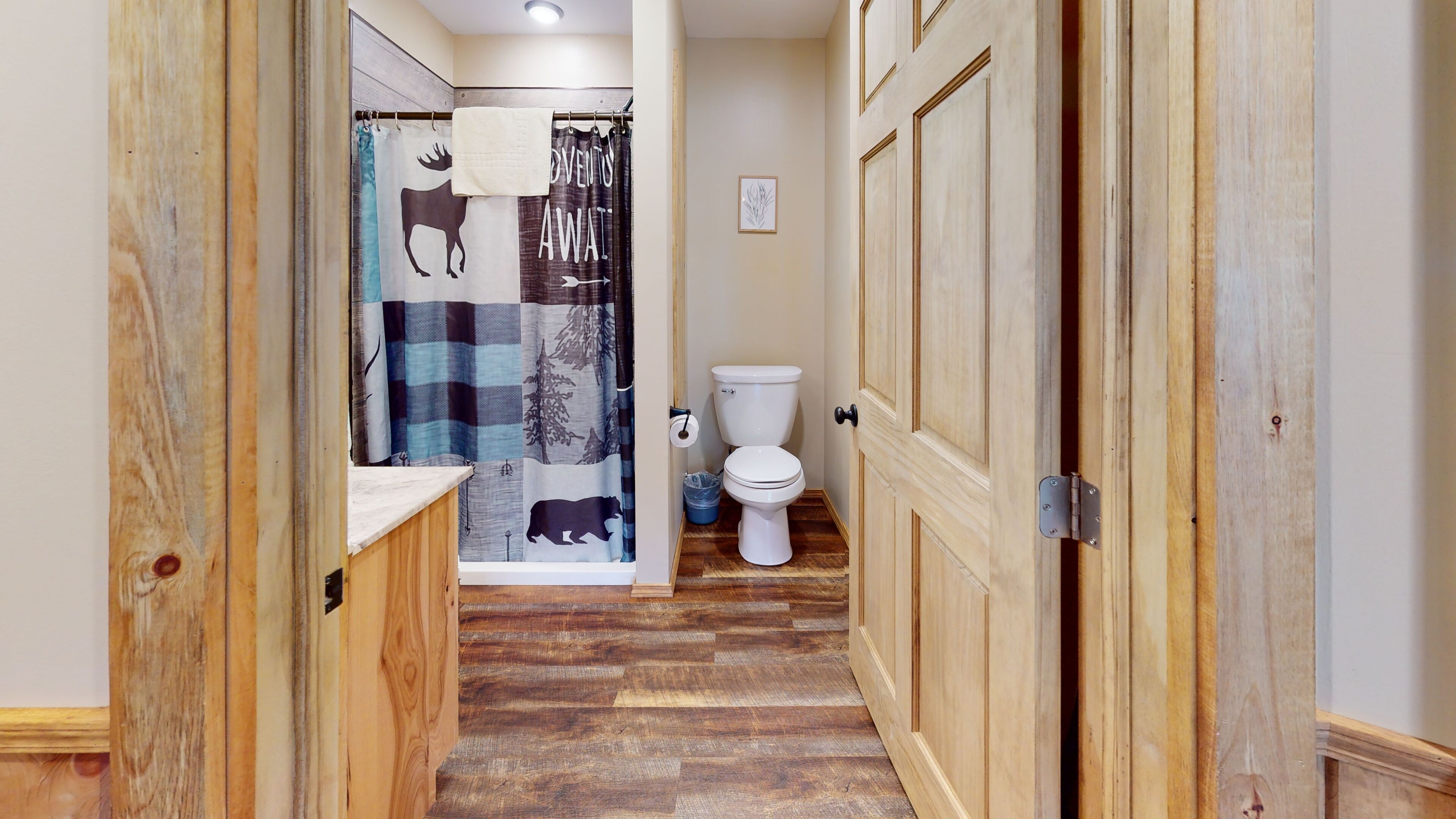 Pasillo con piso de madera que conduce a un baño con inodoro, cortina de ducha con estampados de animales y un cuadro en la pared.