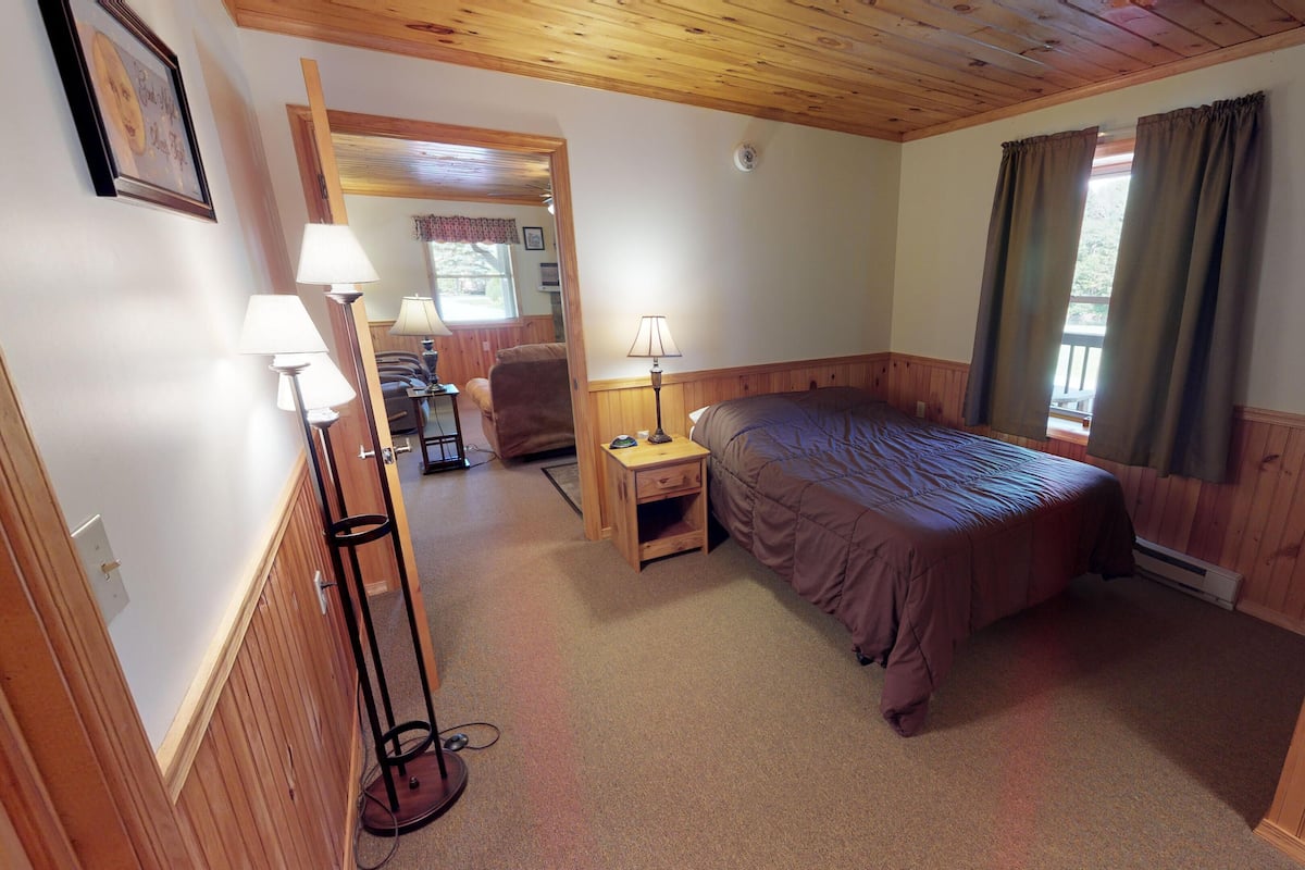 A cozy bedroom with a bed, bedside table, lamp, and a view of the outdoors through a window.
