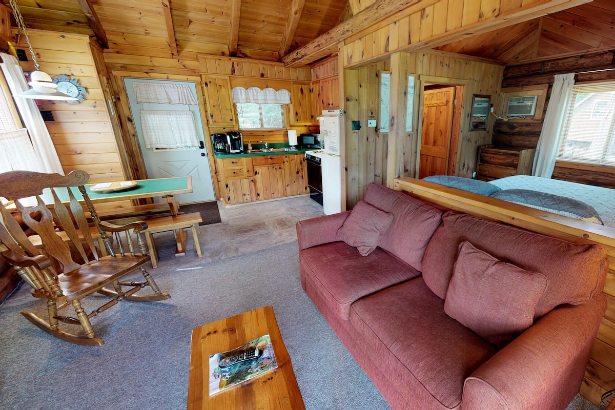 A cozy cabin interior with a wooden dining table, a sofa, a rocking chair, and a kitchenette.