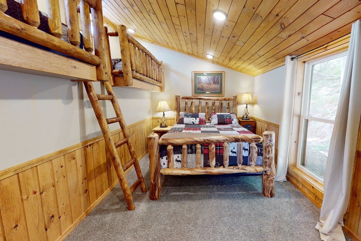 A cozy room with a wooden bunk bed, a ladder, a quilted bedspread, a framed picture on the wall, and a window with curtains.