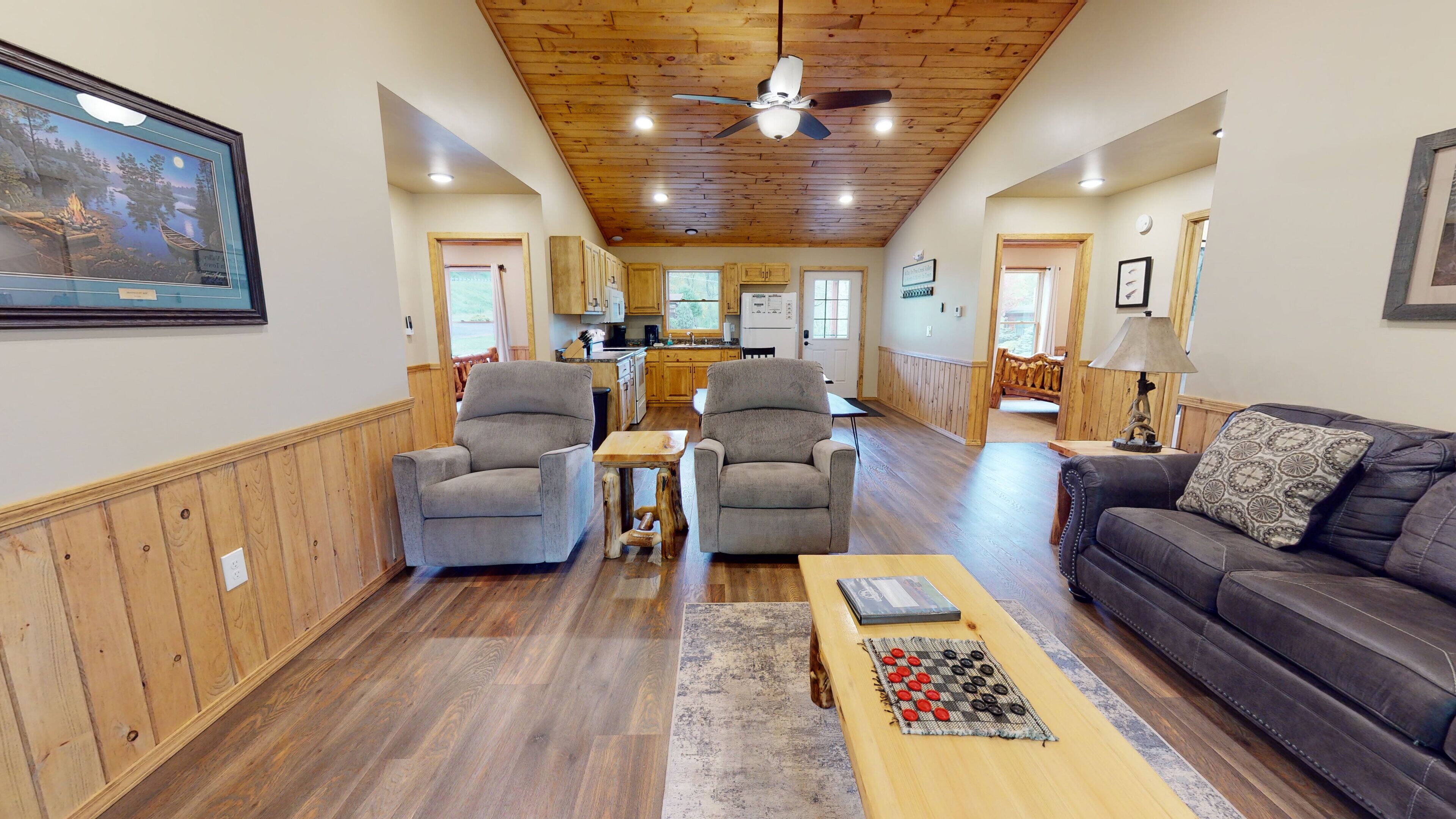A spacious living room with two grey armchairs, a wooden coffee table, and a dark leather sofa. A ceiling fan and recessed lighting are visible.