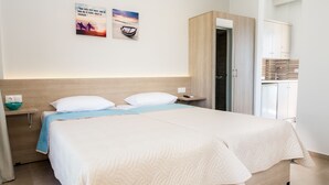 A neatly made bed with a white and light blue bedspread, a bedside table with a bowl, and a wall-mounted picture. - Lola Apartments (Platanias)