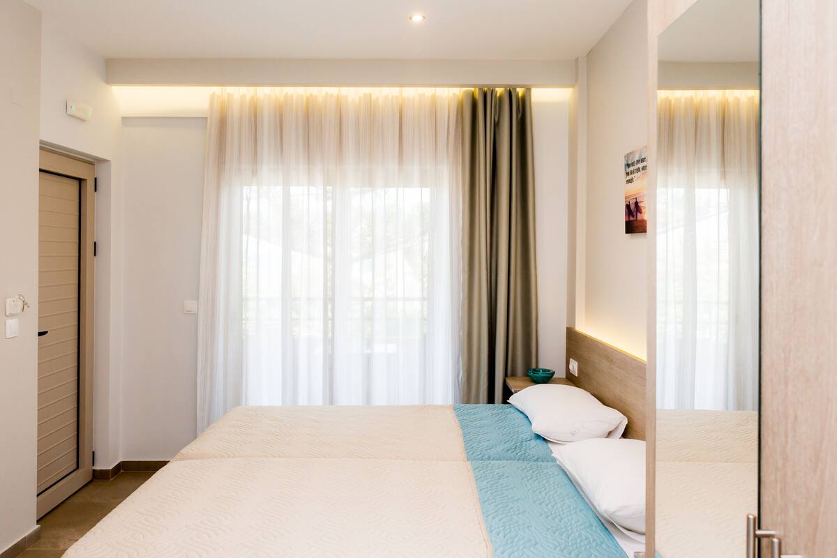 Quarto de hotel com uma cama grande, mesa de cabeceira, espelho e janela com cortinas leves.