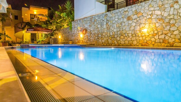 A swimming pool with a stone wall and a building in the background. - Lola Apartments (Platanias)