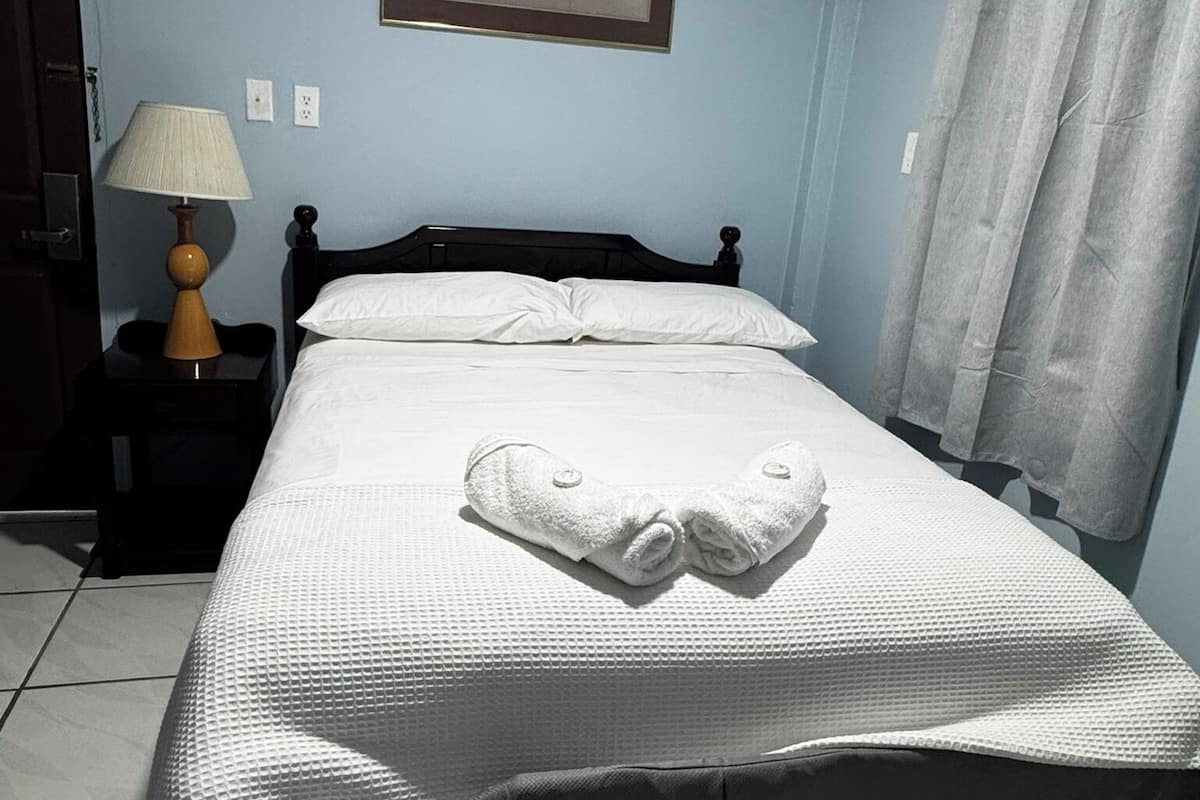 A neatly made bed with a heart-shaped towel, a bedside lamp, and a framed picture on the wall.