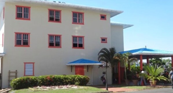 Un edificio de fachada blanca y ventanas con marcos rojos, un área exterior cubierta y un pequeño jardín con una palmera.