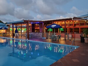 A poolside area with outdoor seating and umbrellas, a covered seating area, and a building with multiple levels and windows.