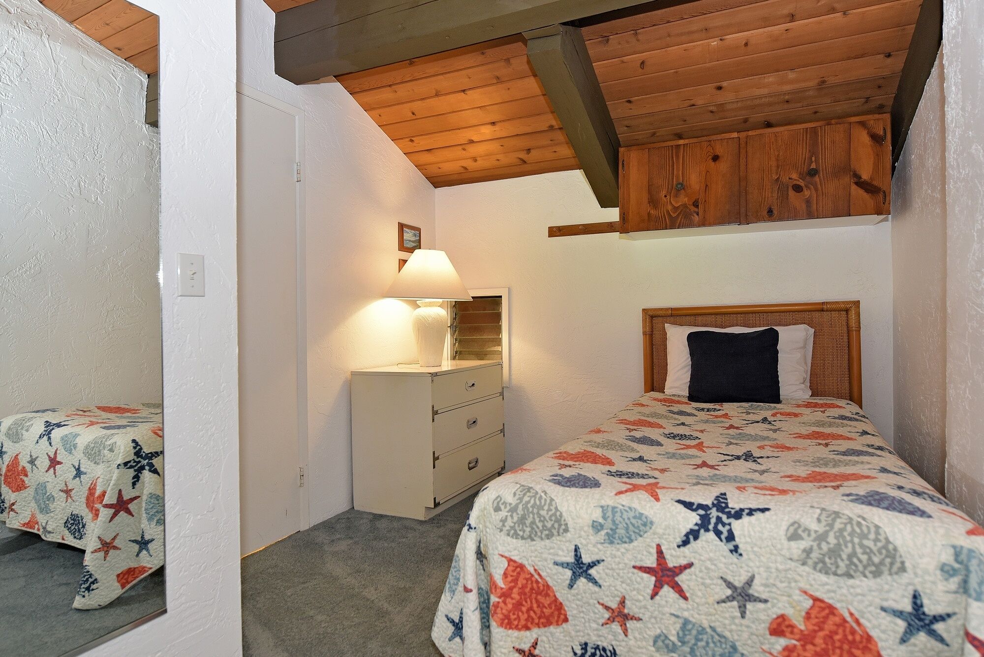 Un dormitorio con techo de madera, una cama con colcha estampada de estrellas, una mesita de noche con lámpara y un aparador.