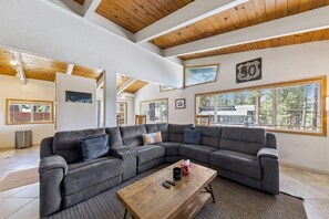 Ein geräumiges Wohnzimmer mit einem großen Ecksofa, einem Holztisch und großen Fenstern mit Blick ins Freie.