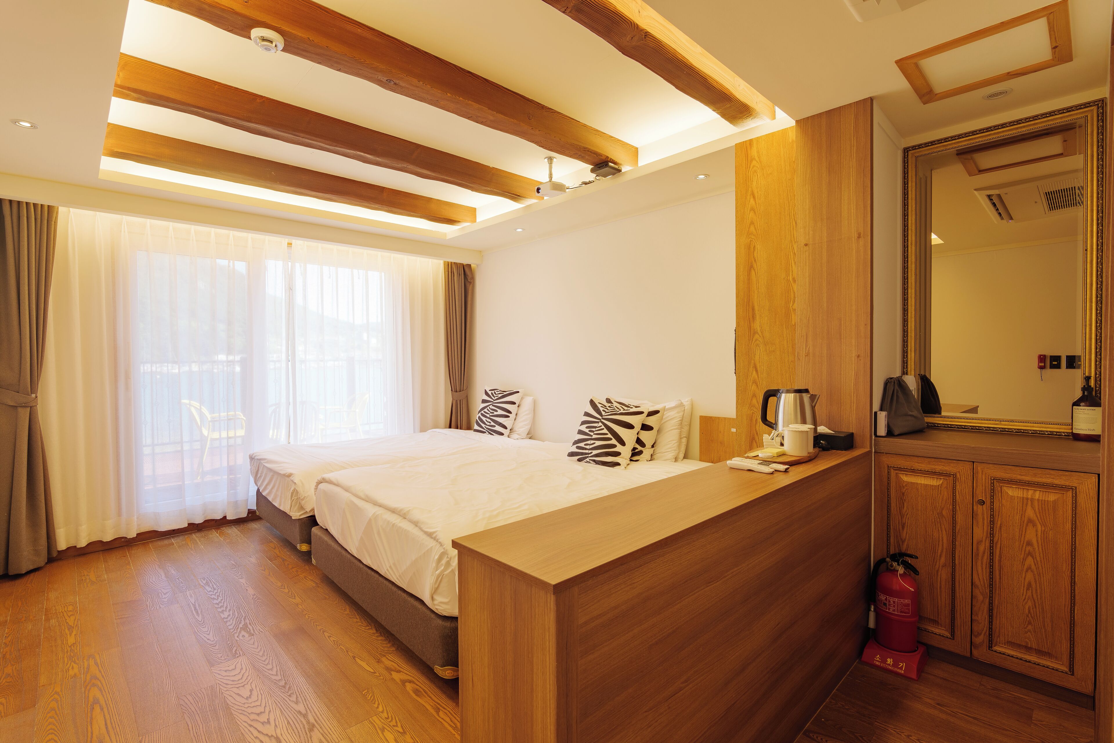 A hotel room with a wooden ceiling, a bed with patterned pillows, a bedside table with a kettle, and a mirror above the door.