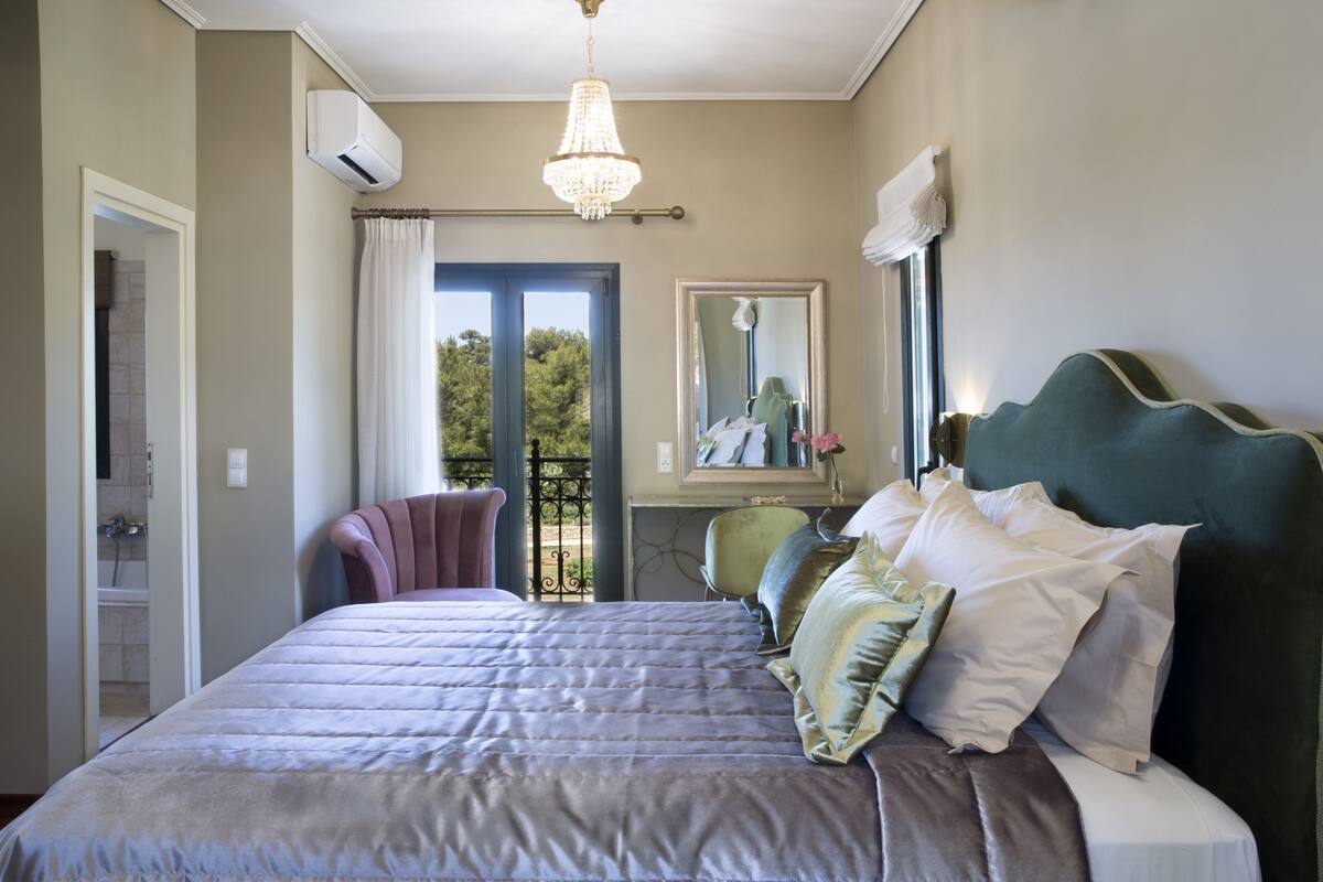 A bedroom with a large bed, a chandelier, a mirror, and a view of the outdoors.