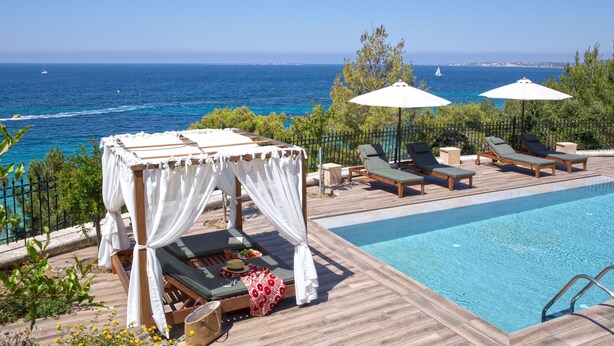 Zona junto a la piscina con terraza de madera, sillas de descanso, una sombrilla y vistas al mar.