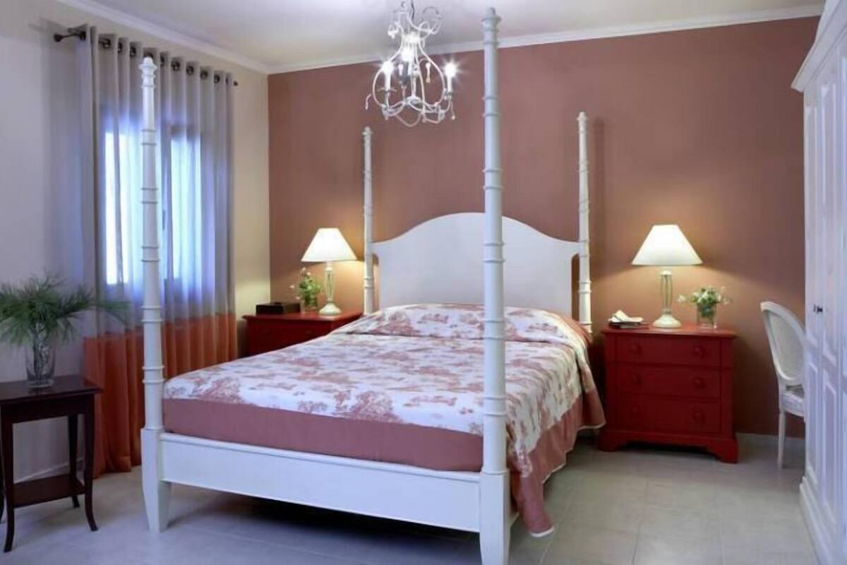 A bedroom with a four-poster bed, two bedside tables with lamps, a chair, and a potted plant.