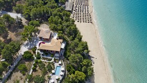 Vista aérea de una propiedad costera con piscina, sombrillas de playa y una fila de sillas de playa.