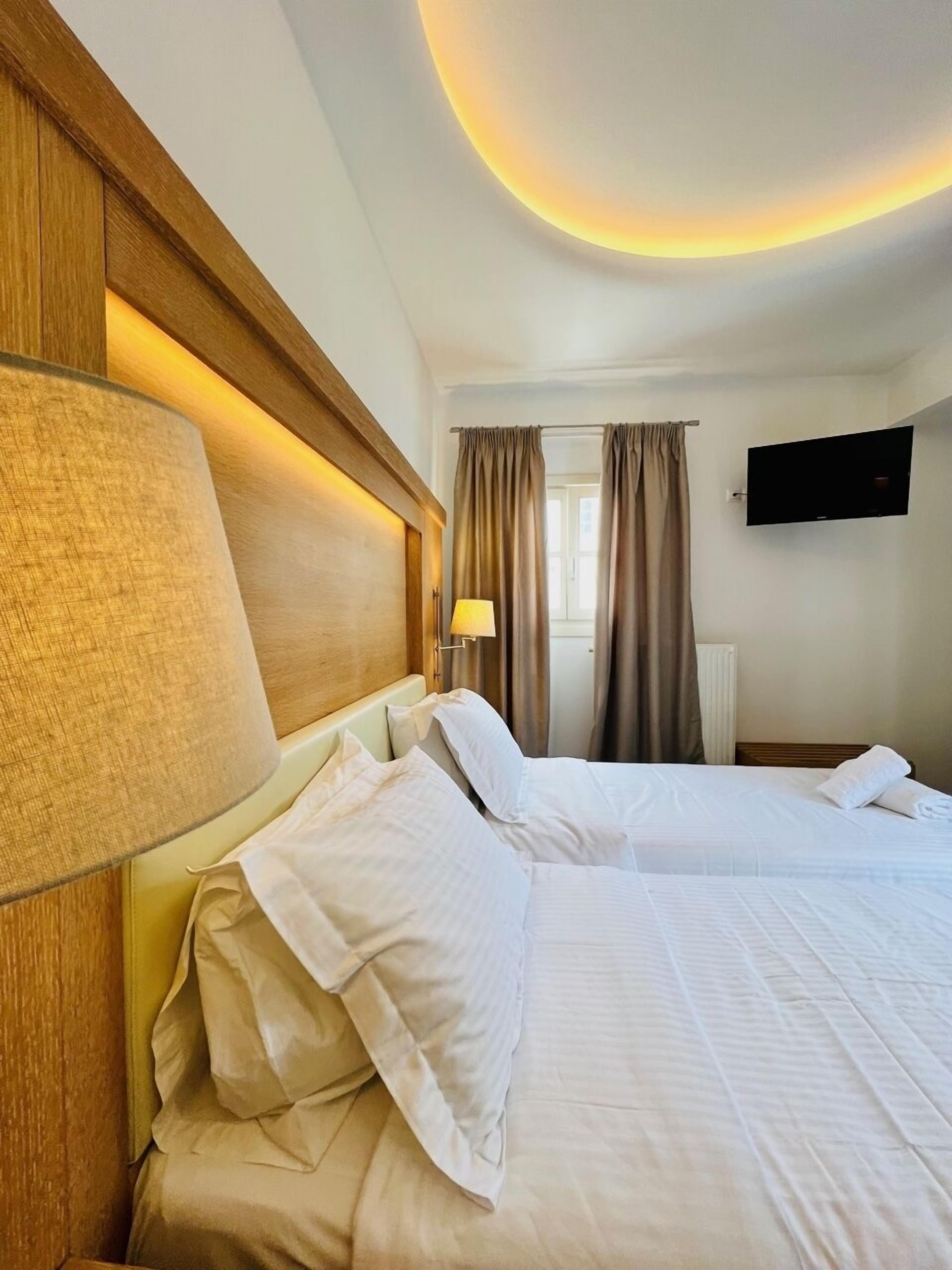 Ein Hotelzimmer mit zwei Betten, einem an der Wand montierten Fernseher und einem Fenster mit Vorhängen.