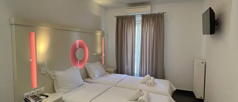 A hotel room with a large bed, a bedside table with a phone and a book, a circular light fixture on the ceiling, and a window with curtains.