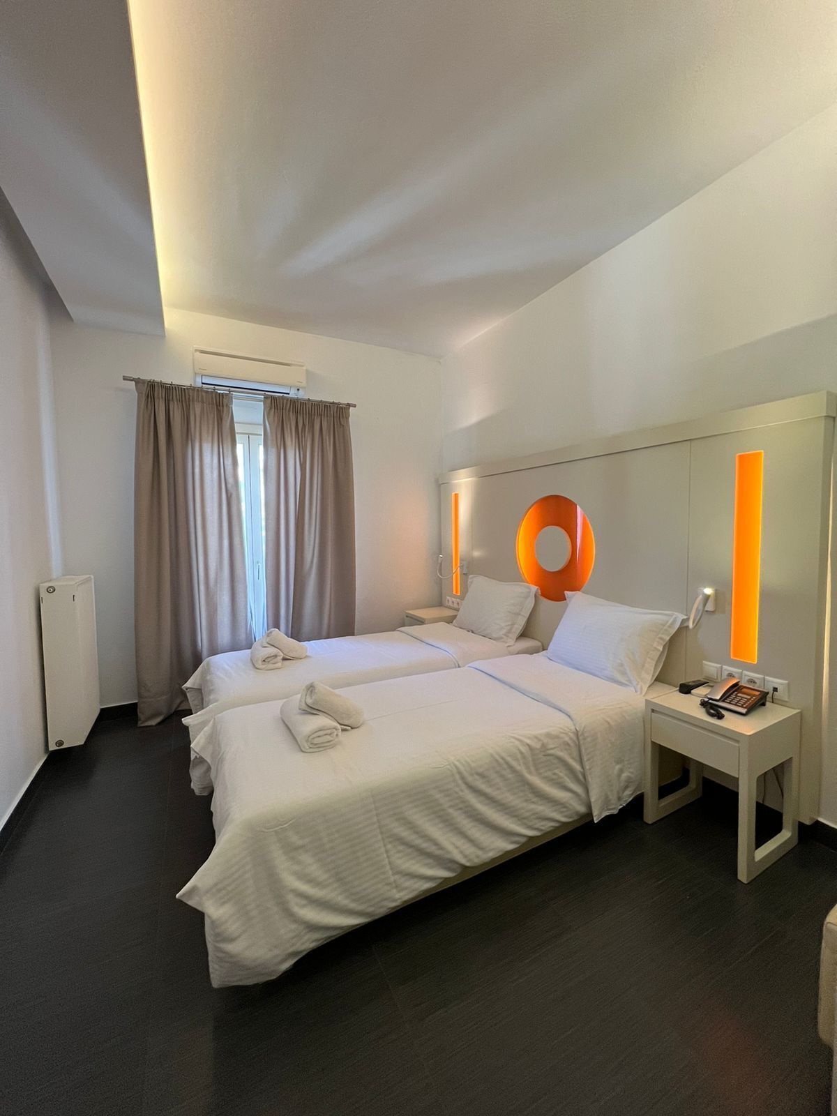 A hotel room with two beds, a bedside table with a phone and a lamp, and a window with curtains.