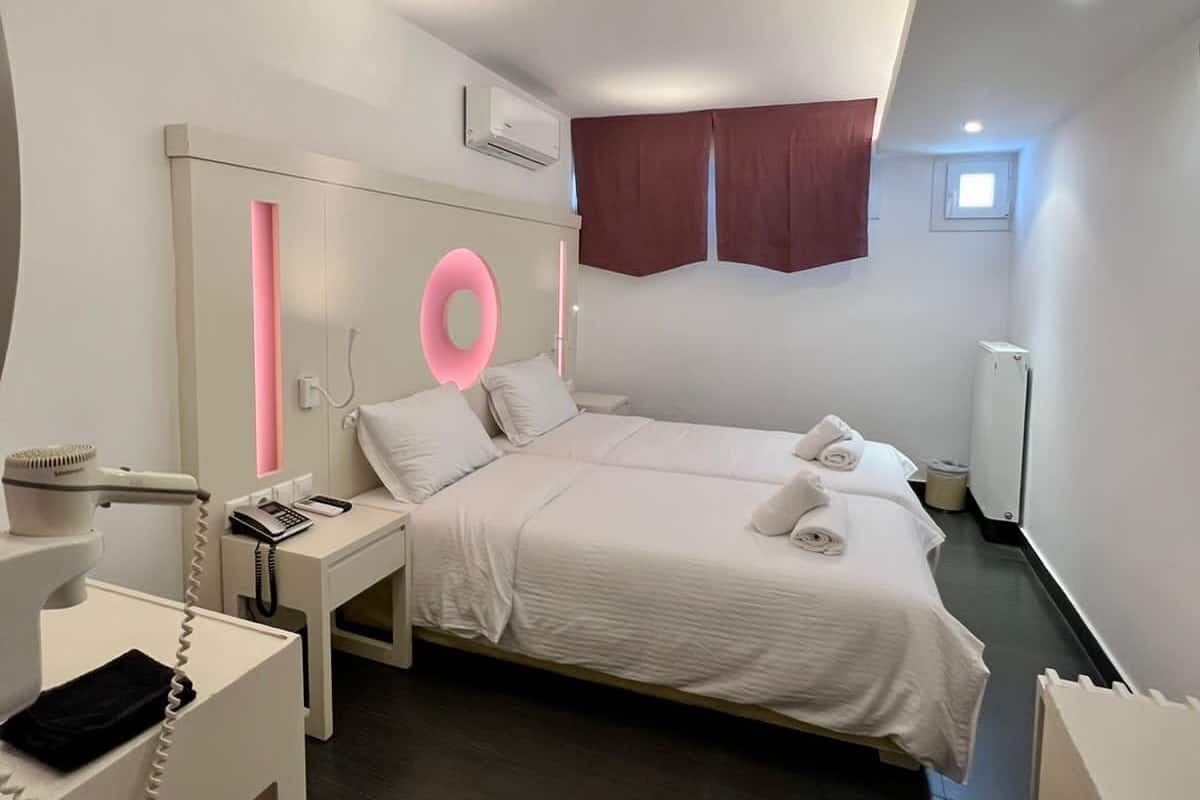 A modern hotel room with a large bed, a bedside table with a phone, a hairdryer, and a mirror. There is a window with curtains and a small skylight.