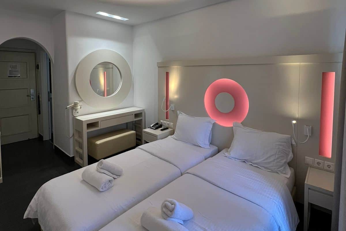 A modern hotel room with two beds, a round mirror, a vanity with a mirror, and a circular light fixture on the ceiling.