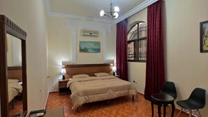 A hotel room with a wooden floor, a bed with a beige duvet, a desk with a vase and a lamp, a chair, and a painting on the wall.