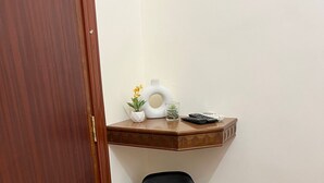 A corner room with a wooden door, a black chair, a small wooden shelf with a vase, a plant, and a glass.