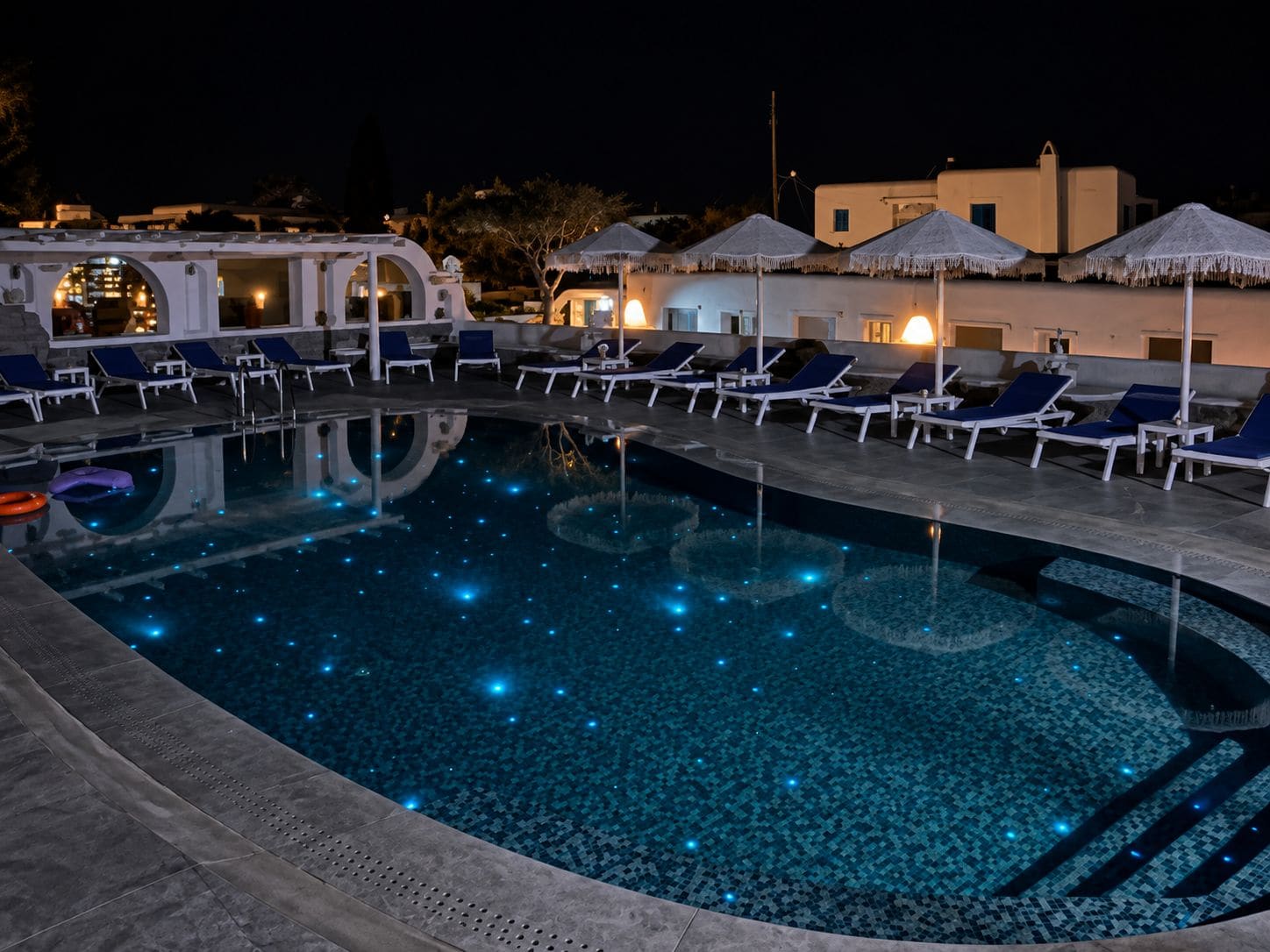 Una piscina con sillas de descanso y sombrillas, rodeada de edificios de noche.
