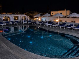 Una piscina con sillas de descanso y sombrillas, rodeada de edificios de noche.