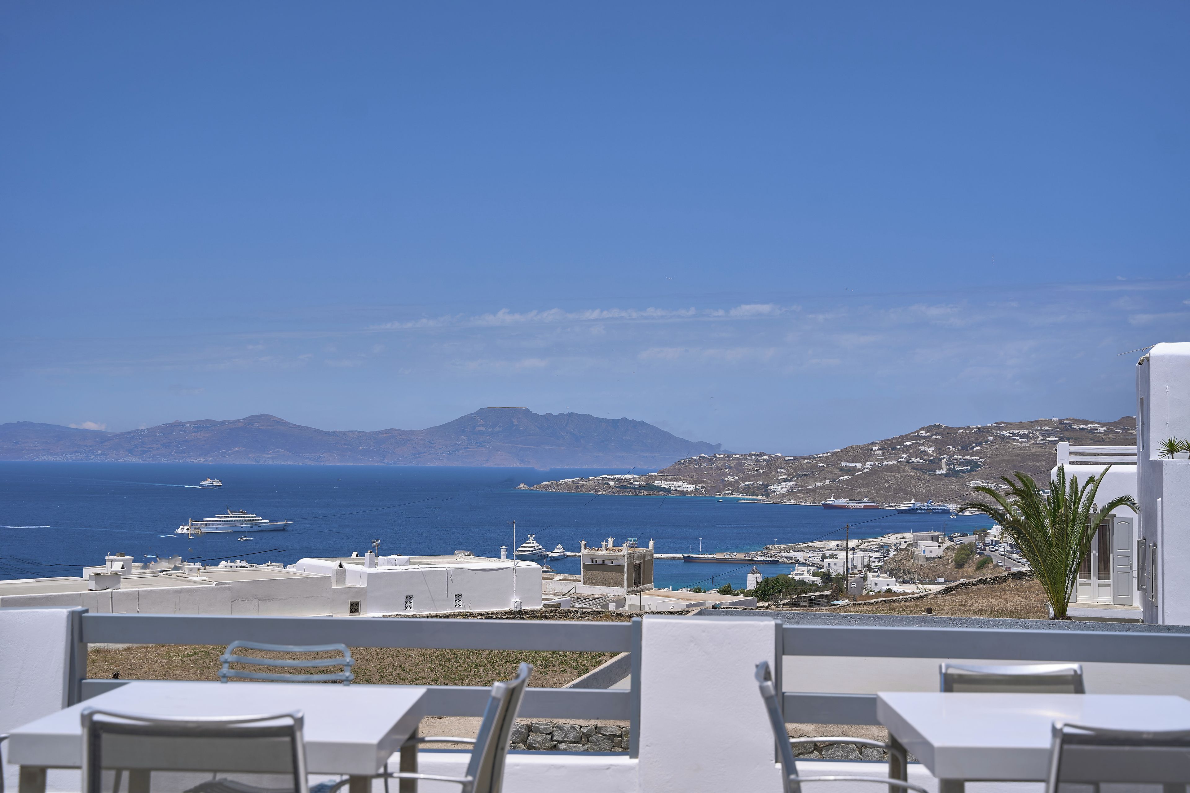 A seaside view from a terrace with white furniture, a clear sky, and a distant coastline with buildings and mountains.