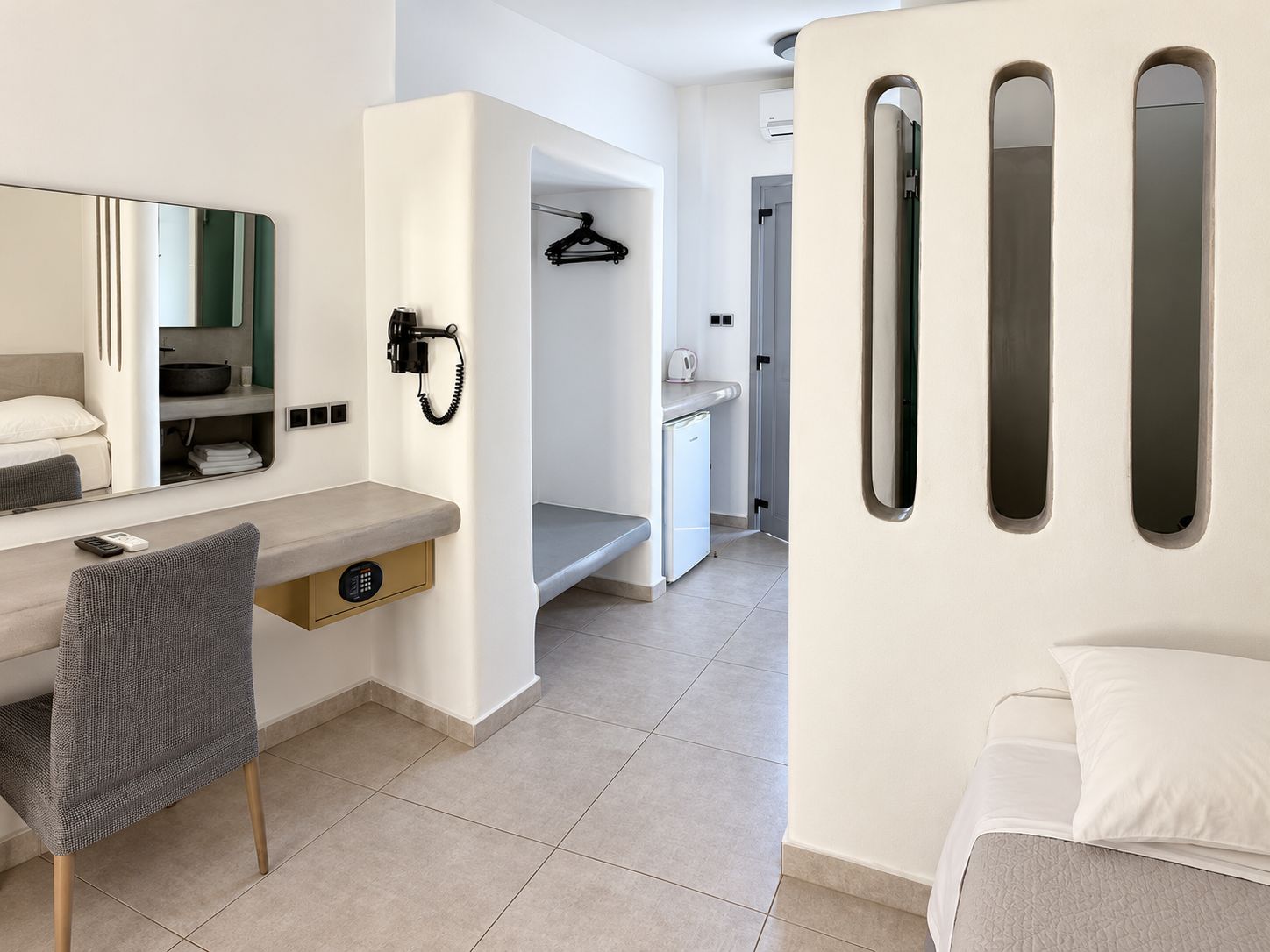 A modern hotel room with a bed, a desk with a chair, a bathroom with a sink and mirror, and a hallway leading to another room.