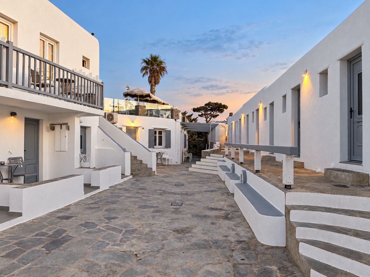 Un patio estrecho empedrado con edificios blancos, una escalera y asientos al aire libre.