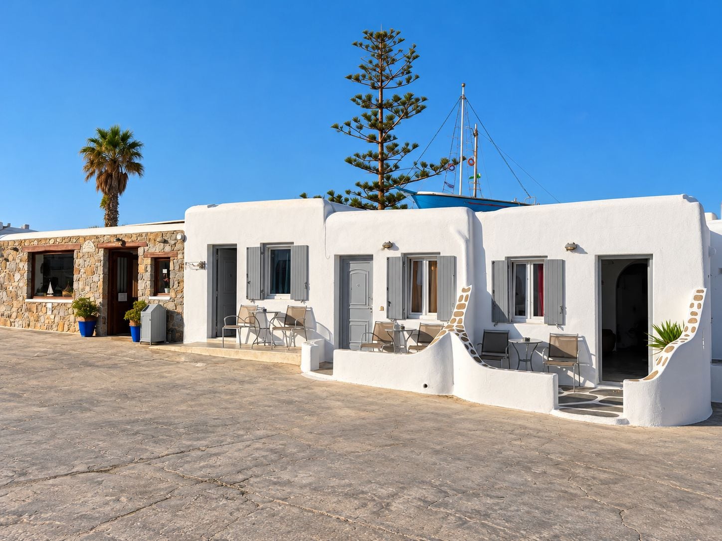 A white building with a stone facade, outdoor seating area, and a palm tree.