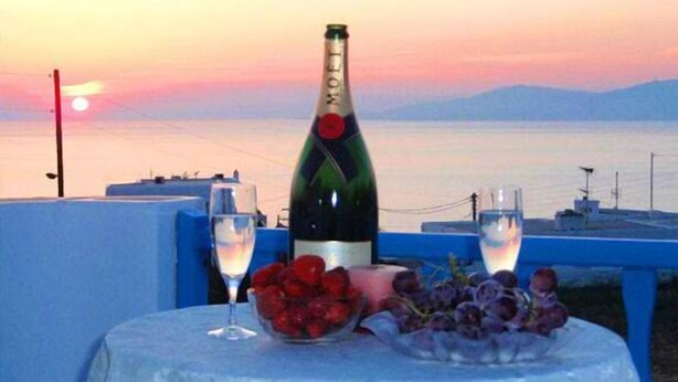 A table with a bottle of wine, two glasses, a bowl of grapes, and a sunset view. - Sofia Village (Mykonos)