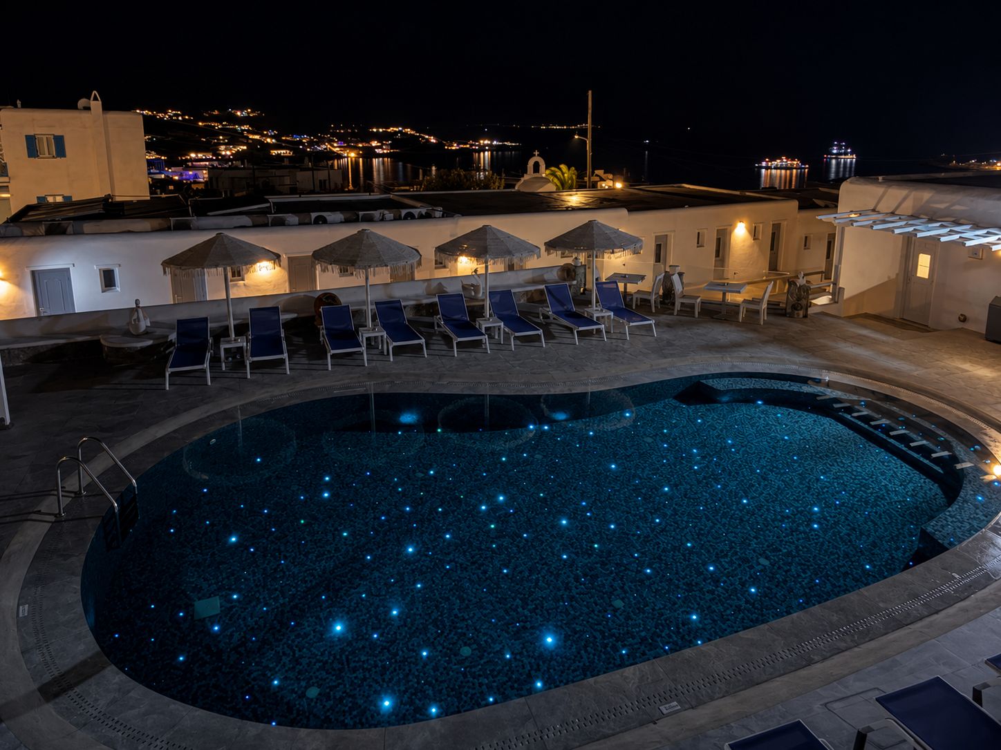 Una piscina nocturna con luces similares a estrellas, sillas de descanso y sombrillas.