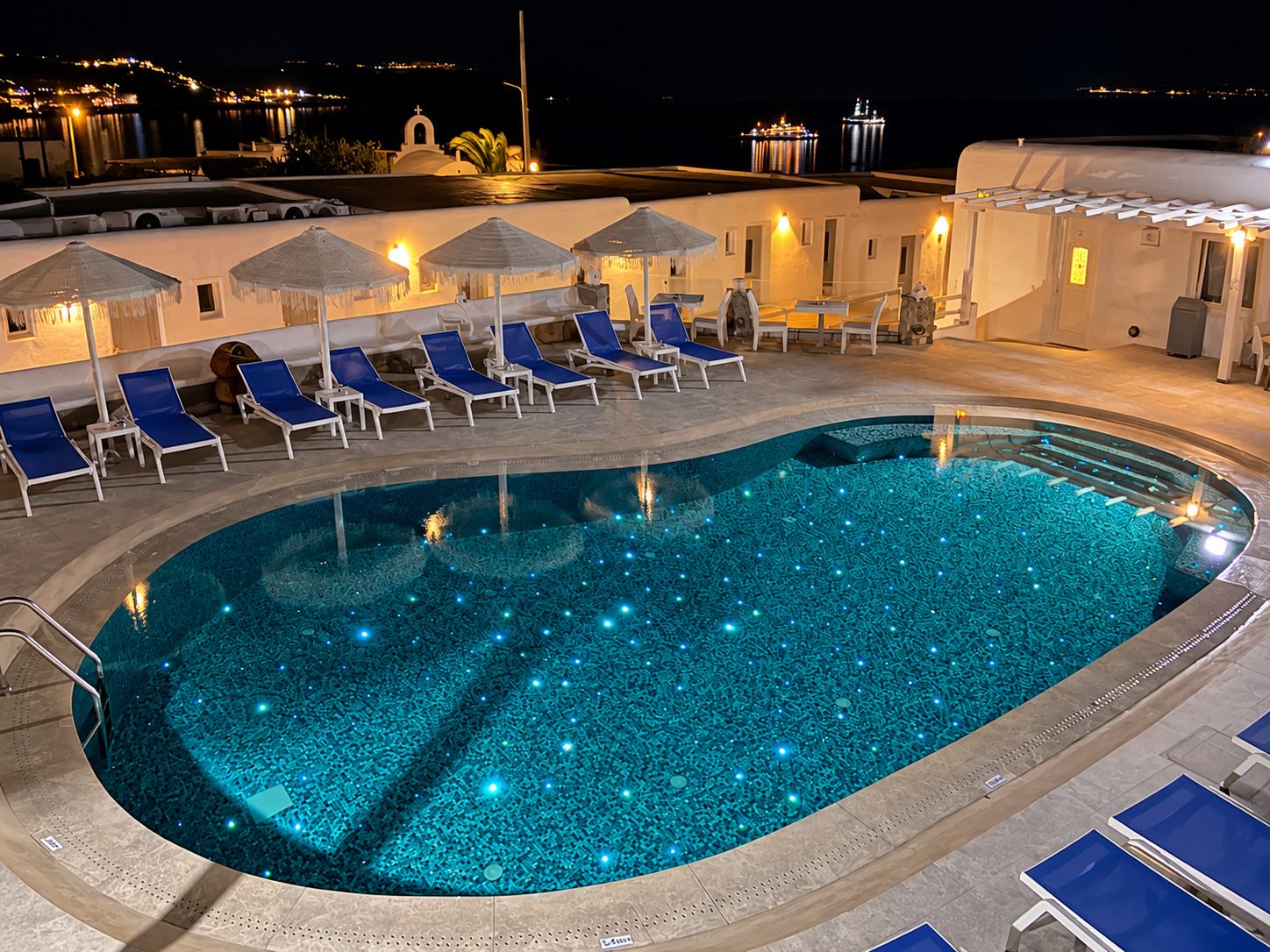 Una piscina con sillas de descanso y sombrillas, rodeada por un edificio con iluminación exterior.