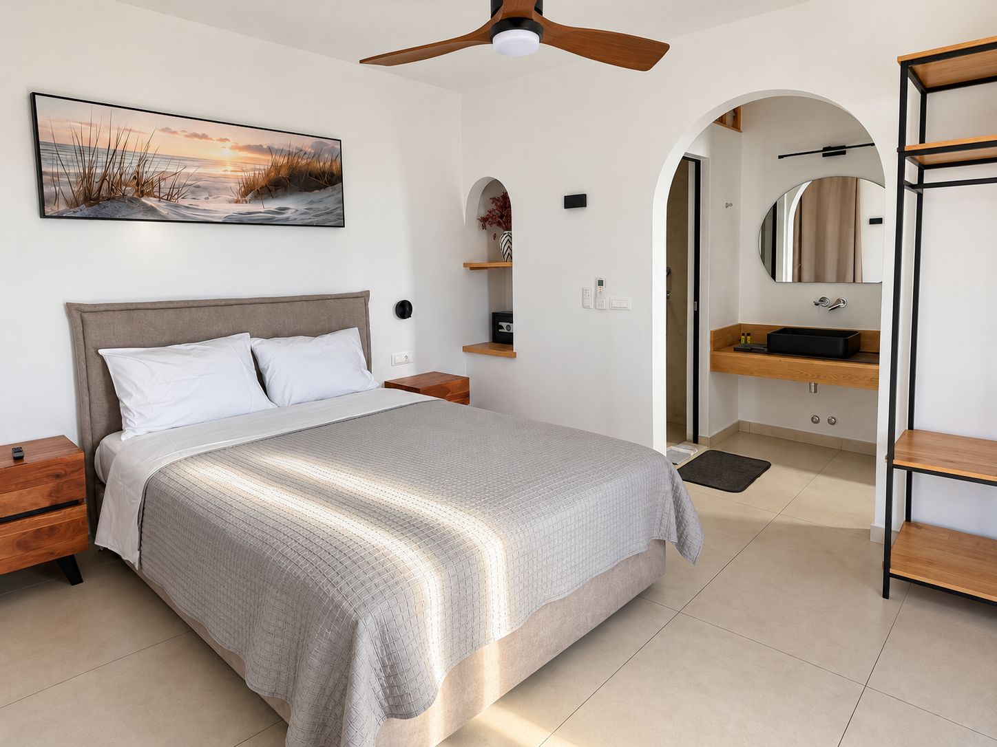 A modern bedroom with a bed, a ceiling fan, a painting, a bathroom with a sink and mirror, and a wooden shelf.