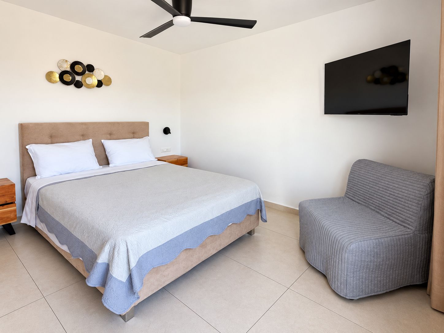 A bedroom with a bed, a chair, a ceiling fan, a wall-mounted TV, and a decorative wall piece.