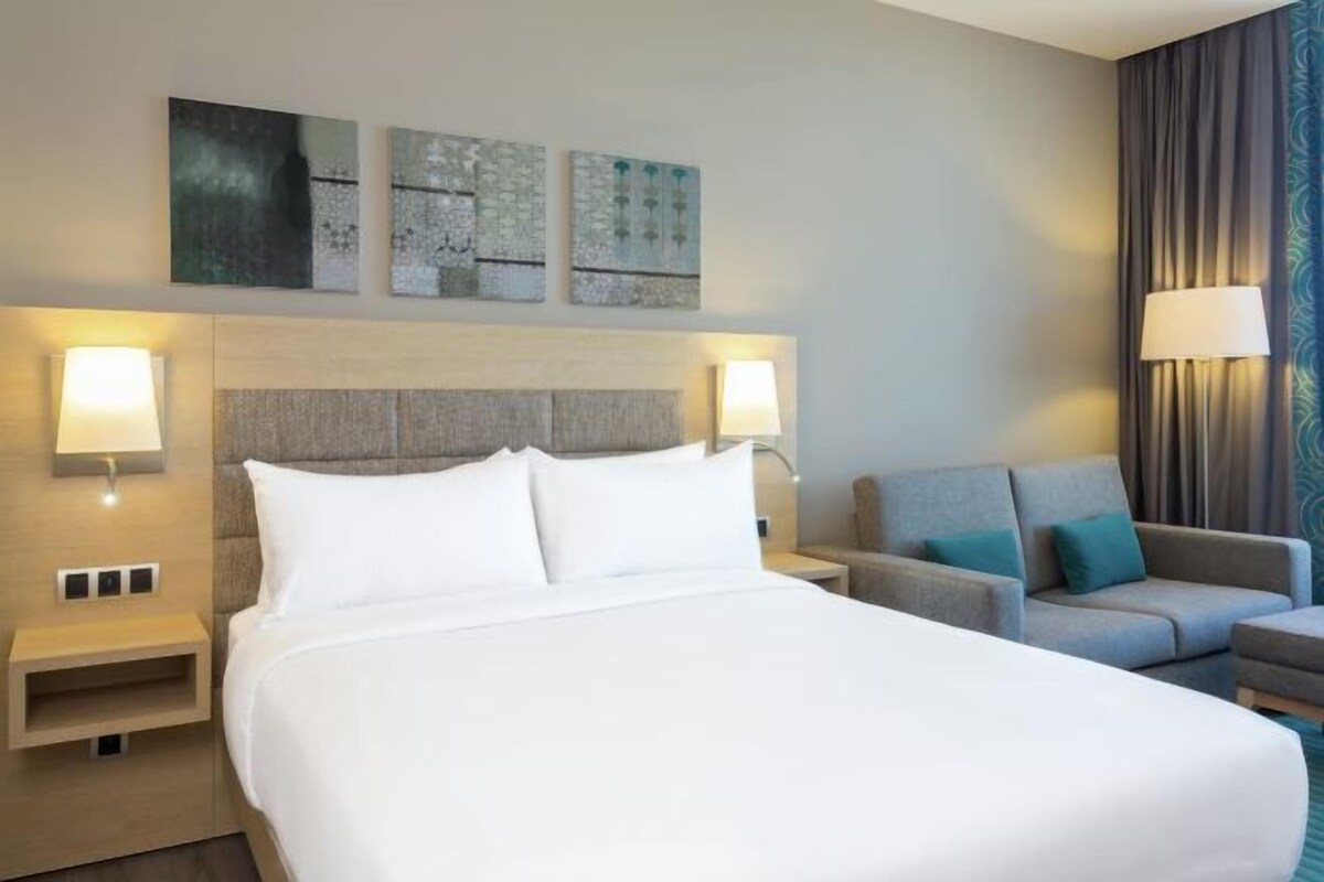 A hotel room with a large bed, two bedside tables with lamps, a grey sofa, and artwork on the wall.