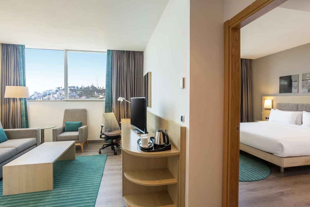 A hotel room with a bed, a desk with a computer, a chair, a sofa, and a view of the city.