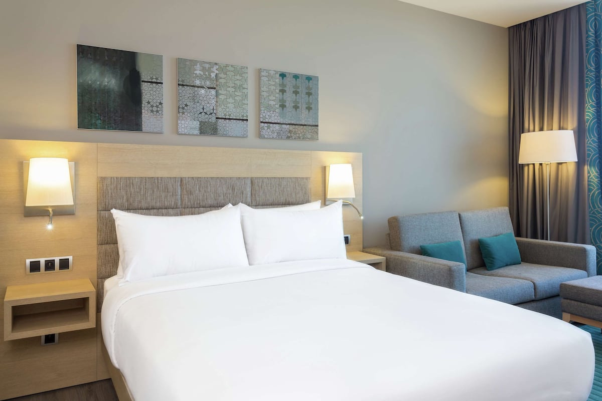 A hotel room with a large bed, two bedside tables with lamps, a grey sofa, and abstract wall art.