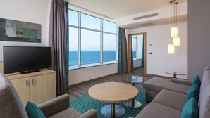 A modern hotel room with a large window offering a sea view, a flat-screen TV on a wooden cabinet, a round glass table, and a sofa with teal cushions. - Hilton Garden Inn Tanger City Center (Tangier)